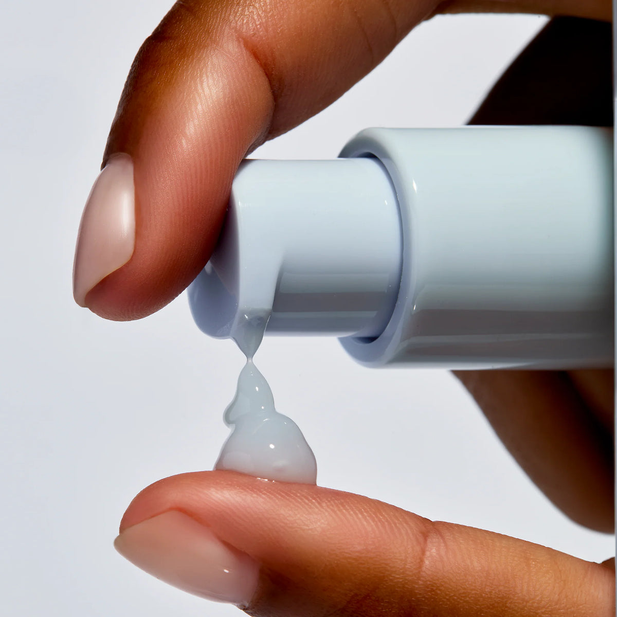 Hand holding a blue bottle with a droplet of liquid being squeezed out on a light gray background